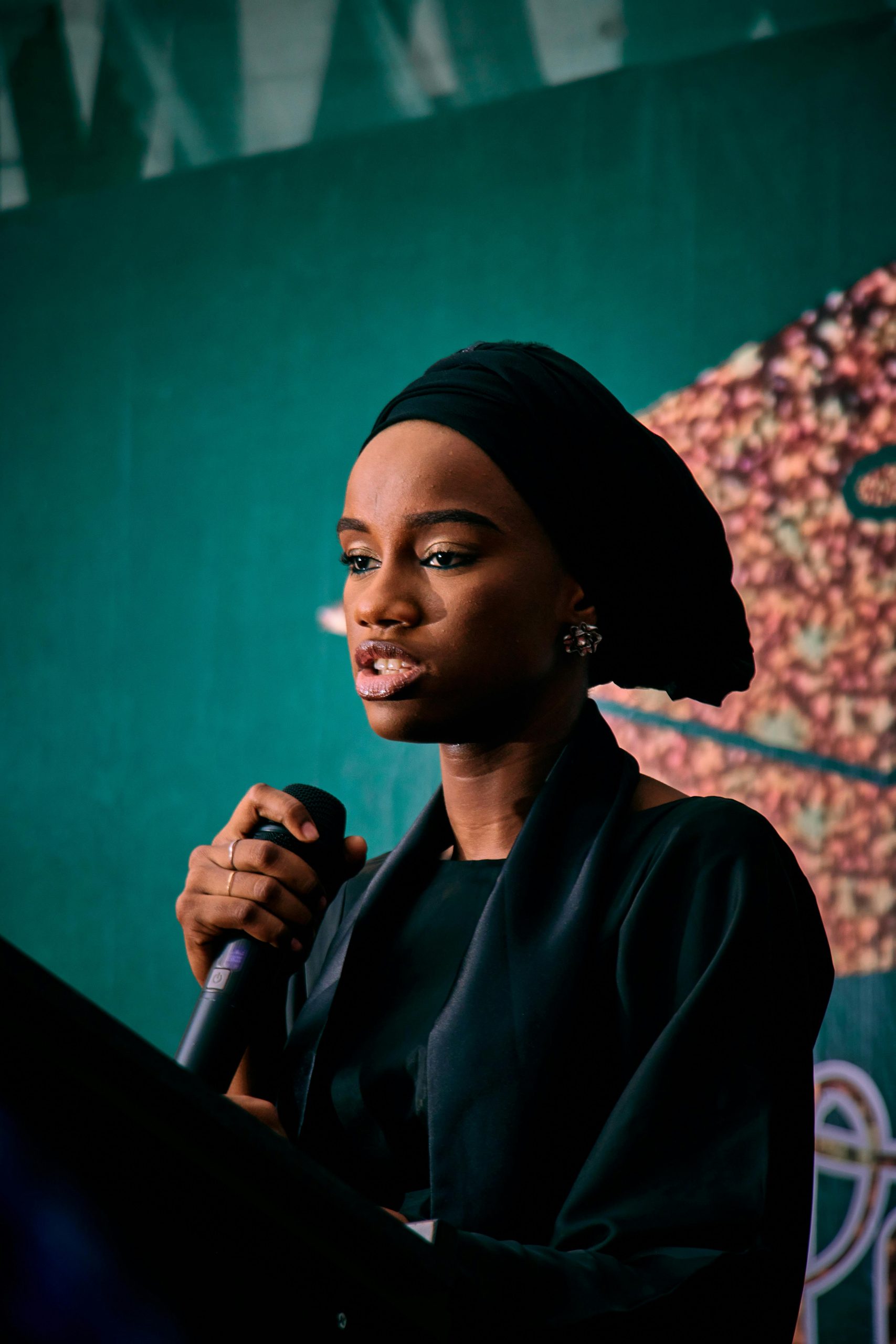 A confident woman in a headscarf speaks passionately at a podium, microphone in hand.